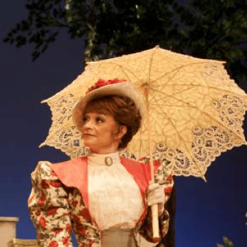 A performer playing Mrs Warren on stage in Victorian-style costume, holding a white parasol