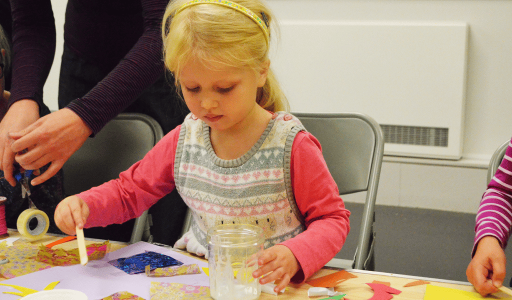 A young blonde girl partakes in craft activities with a look of concentration on her face