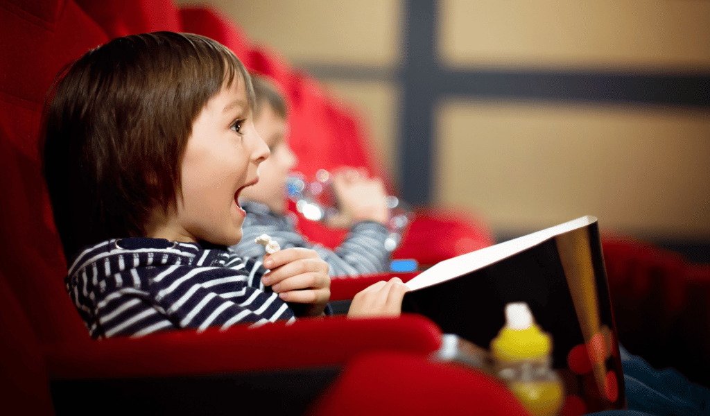 a young child sits in a theatre chair with popcorn with a look of wonder on their face