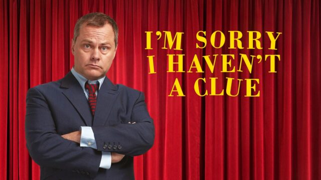 I'm Sorry I Haven't A Clue hero image featuring Jack Dee wearing a blue suit, standing with his arms folded and looking grumpy and confused