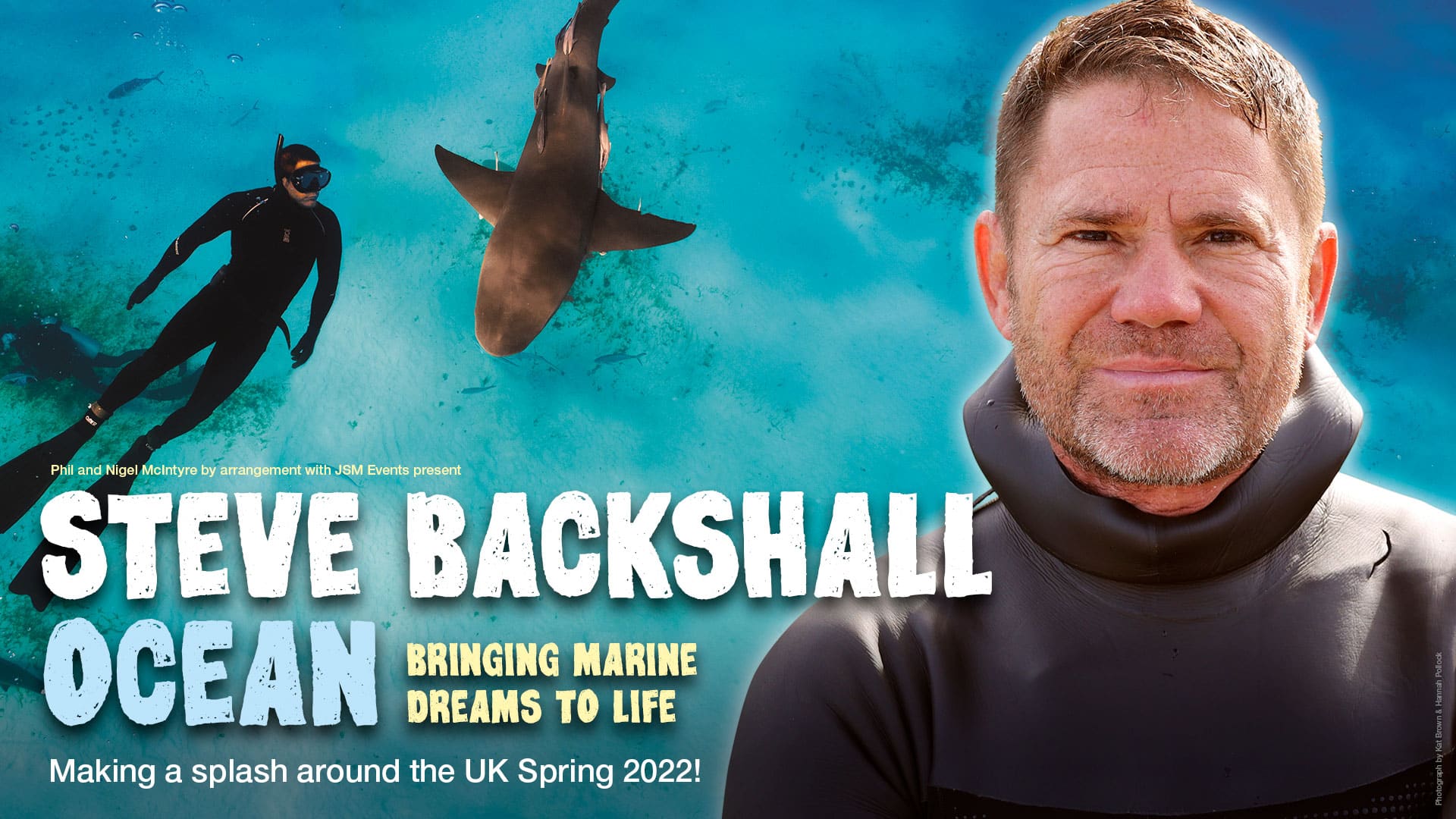 A close up of Steve Backshall. In the background, you can see a shark in the ocean and a diver swimming nearby