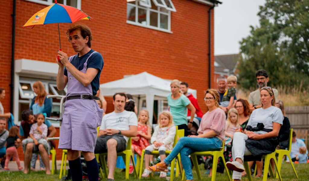 Newcourt Community Festival 2021 Donald Craigie performing in front of families. He's got a colourful umbrella and is wearing purple dungarees
