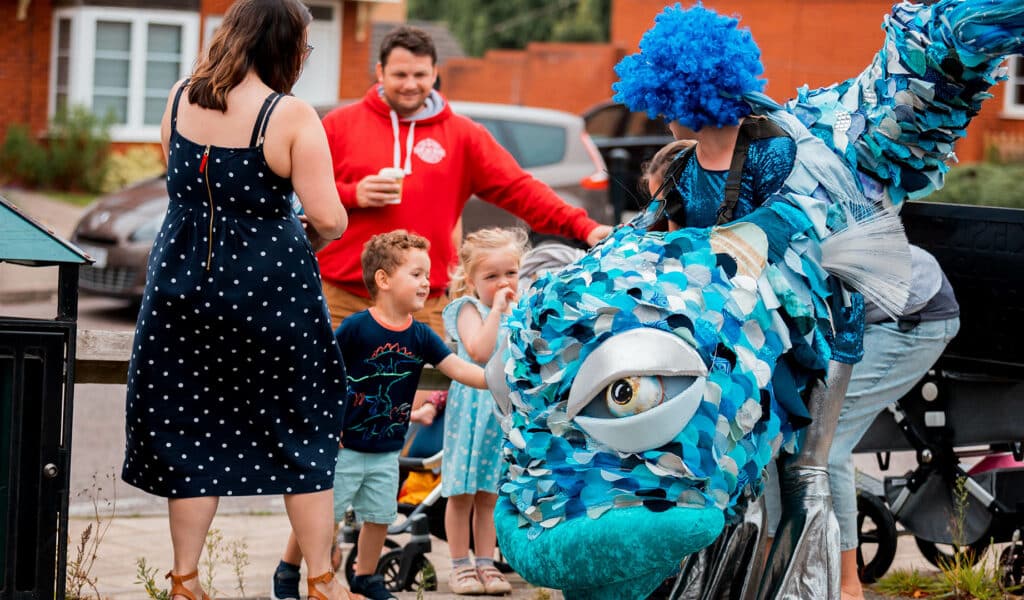 Newcourt Community Festival 2021 A young family is having a closer look at a big blue fish puppet, carried by a performer in a bright blue wig!