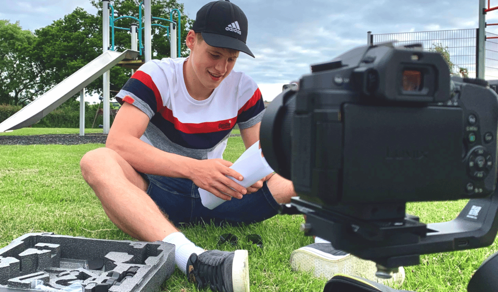 A young man sits on the grass in a park, holding a script and setting up filming equipment