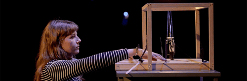A woman reaches into a wooden cube, which has a small carboard cutout figures inside