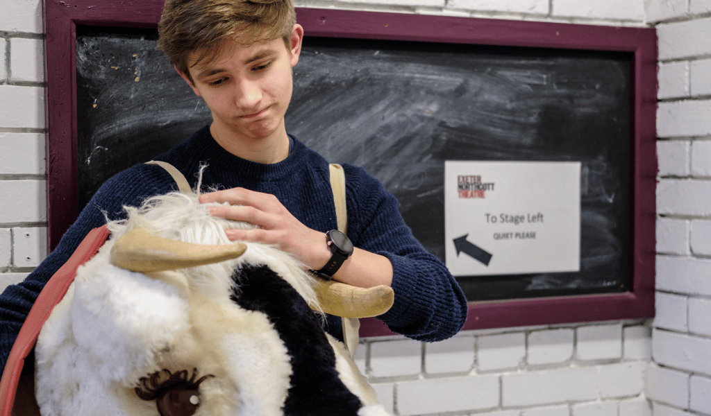 A young man looks down at a fake cow head – to their right, a sign saying ‘stage left, quiet please’