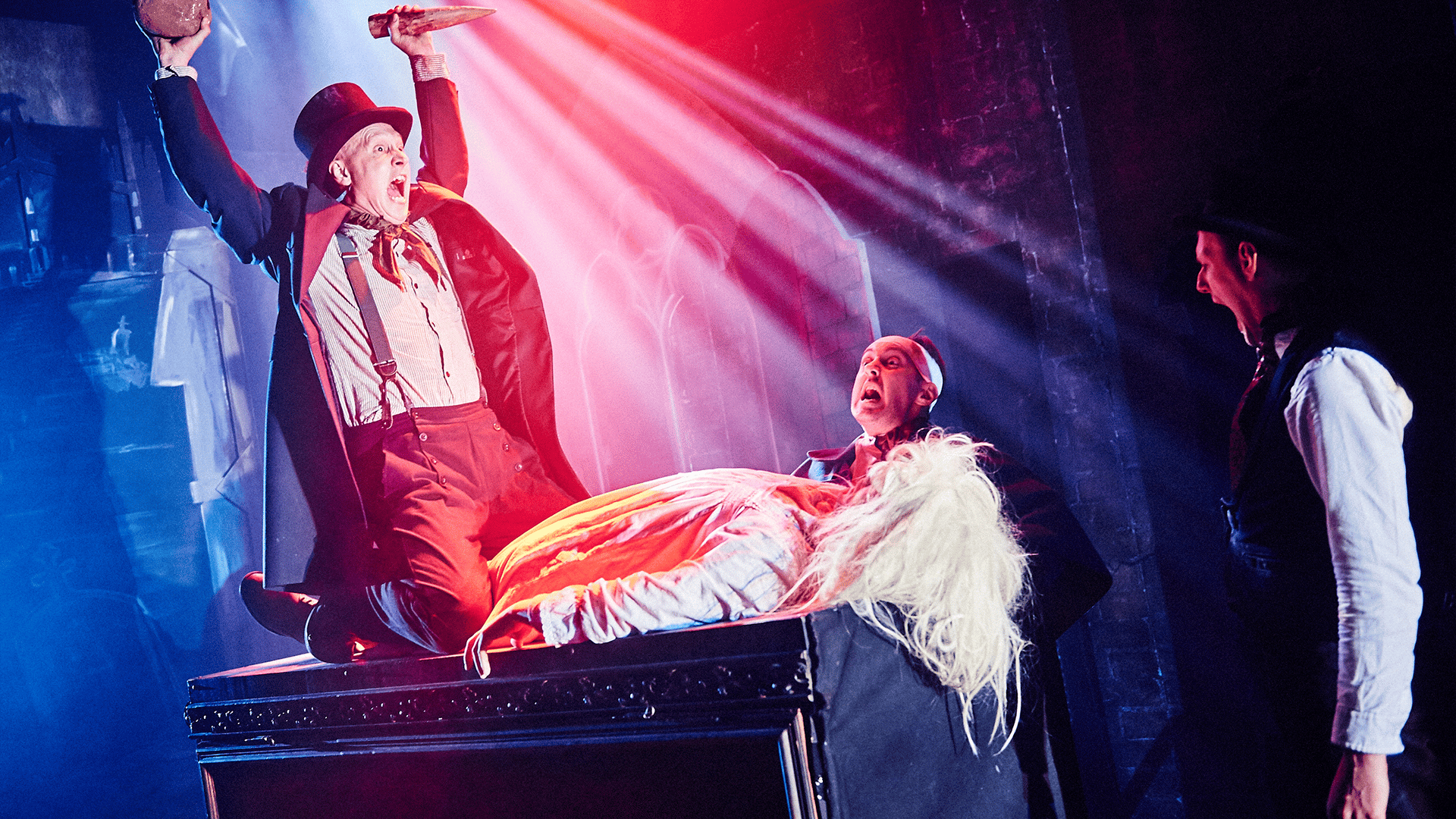 Inside a church. On the left-hand side, a performer dressed in victorian clothing and wearing a top hat holds two wooden stakes in the air whilse kneeling on another perfromer dressed in white with a blond wig. Both of them are on a coffin. In the centre of the image, a performer looks up, mouth gaping-open. To the right-hand side of the image, a perfomer looks to the left hand side, mouth gaping-open.