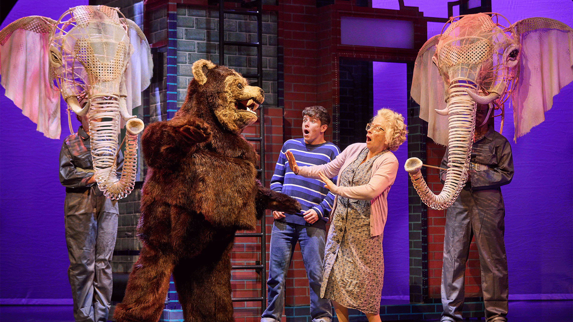 A young man and an older woman are startled by a large grizzly bear puppet. Two elephant puppets stand either side of this group.