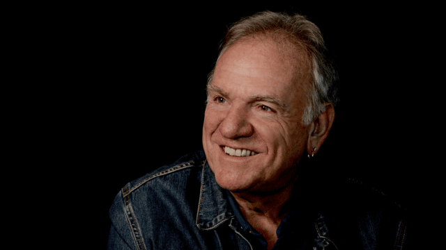 Black background - on the right hand side, Ralph McTell, looking ot the left side, wearing a dark denim jacket and smiling.