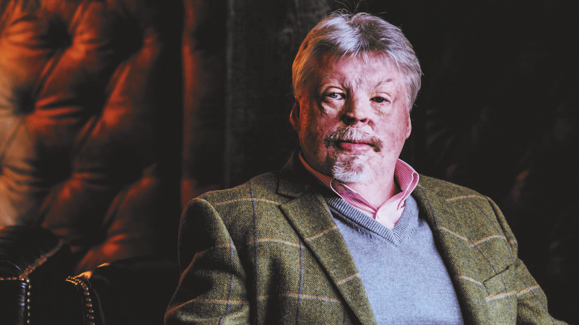 Right-hand side, Simon Weston, sitting in front of a leather chair, wearing a shirt, jumper and tweed jacket, looking to the left side