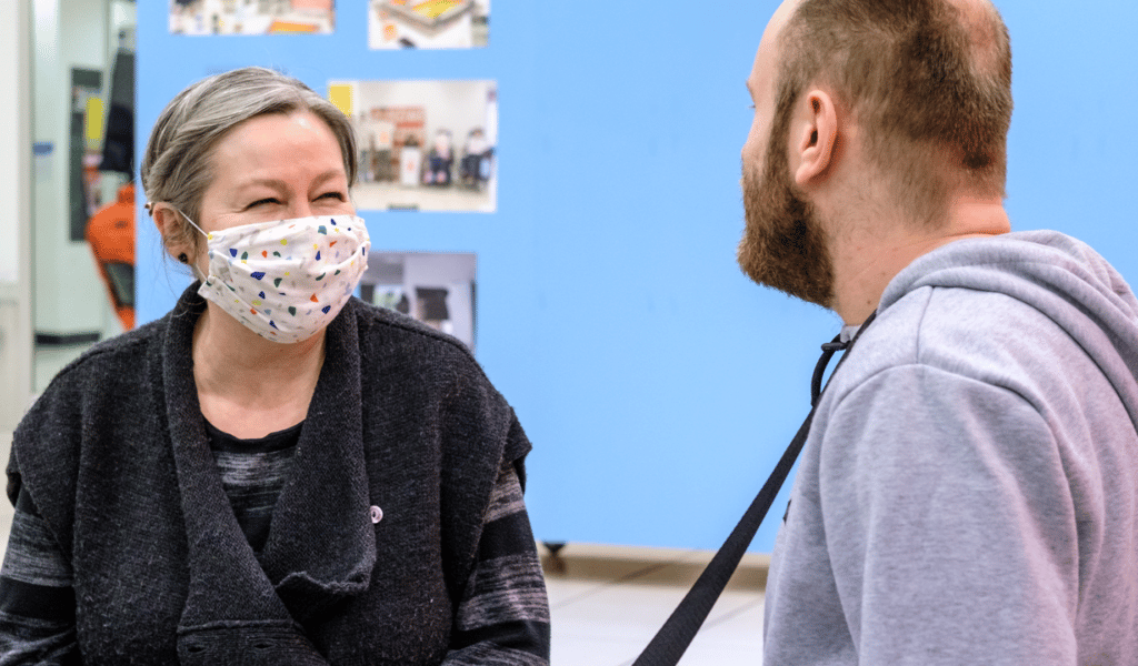Lisa Hudson, Producer for Children and Young People at Exeter Northcott Theatre, wearing a dark showel and a confetti-graphic face mask, smiles at Adam, a Creative Connections member.