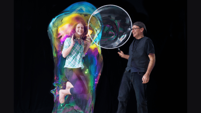 The Amazing Bubble Man on stage with a girl, who is jumping up, completely surrounded by a giant, colourful bubble