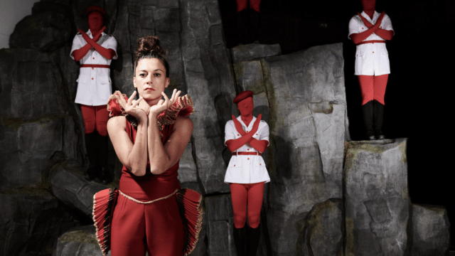 A female dancer in red costume is looking straight at the camera, supporting her chin with her hands. Strange figures are mimicking her in the background.